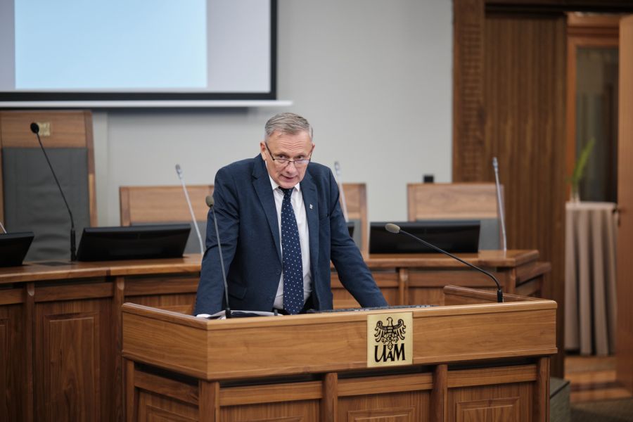 Mężczyzna w garniturze przemawia przy mównicy z logo Uniwersytetu im. Adama Mickiewicza w Poznaniu. W tle widoczny fragment ekranu prezentacyjnego.