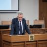 Mężczyzna w garniturze przemawia przy mównicy z logo Uniwersytetu im. Adama Mickiewicza w Poznaniu. W tle widoczny fragment ekranu prezentacyjnego.