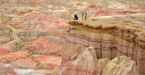 Trzy osoby stojące na krawędzi skalnego klifu, z widokiem na rozległy, kolorowy krajobraz wzgórz i formacji skalnych.