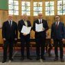 Cztery osoby stoją w reprezentacyjnym wnętrzu i prezentują podpisane dokumenty po oficjalnym spotkaniu, na tle przeszklonych okien i drewnianych ścian.