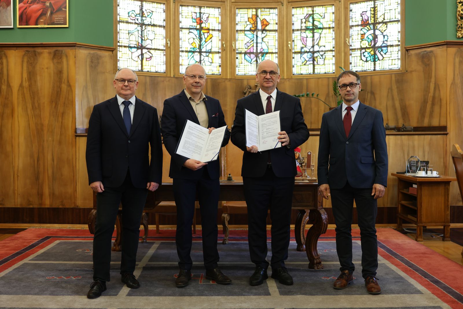 Cztery osoby stoją w reprezentacyjnym wnętrzu i prezentują podpisane dokumenty po oficjalnym spotkaniu, na tle przeszklonych okien i drewnianych ścian.