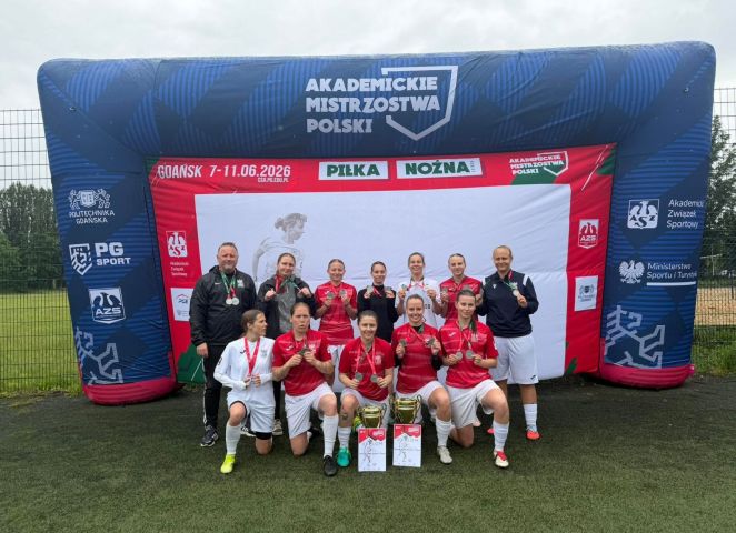 Drużyna piłkarska pozuje do zdjęcia i prezentuje medale oraz puchary podczas Akademickich Mistrzostw Polski w Gdańsku; w tle widoczna jest dmuchana brama z napisem „Akademickie Mistrzostwa Polski – Piłka Nożna”.