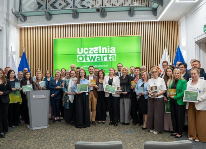 Duża grupa osób stoi w sali konferencyjnej i pozuje do wspólnego zdjęcia, trzymając dyplomy i statuetki; w tle widoczny jest ekran z napisem „uczelnia otwarta” oraz flagi i elementy wnętrza.