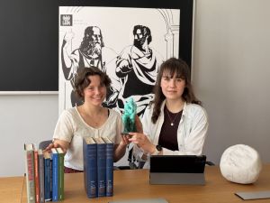 Dwie młode kobiety siedzą przy stole w sali Wydziału Filozoficznego UAM. Wspólnie prezentują niewielką zieloną figurkę.