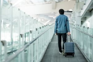  Person walking through a modern glass corridor, pulling a small wheeled suitcase. Dressed casually in a denim jacket, black pants, and white shoes. The setting resembles an airport or transit hub, with a carpeted floor, high ceiling, and visible structural beams—evoking themes of travel, mobility, and transition.