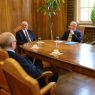 Grupa osób siedzi przy dużym drewnianym stole w eleganckiej sali i rozmawia podczas spotkania, na stole znajdują się filiżanki i szklanki z wodą.