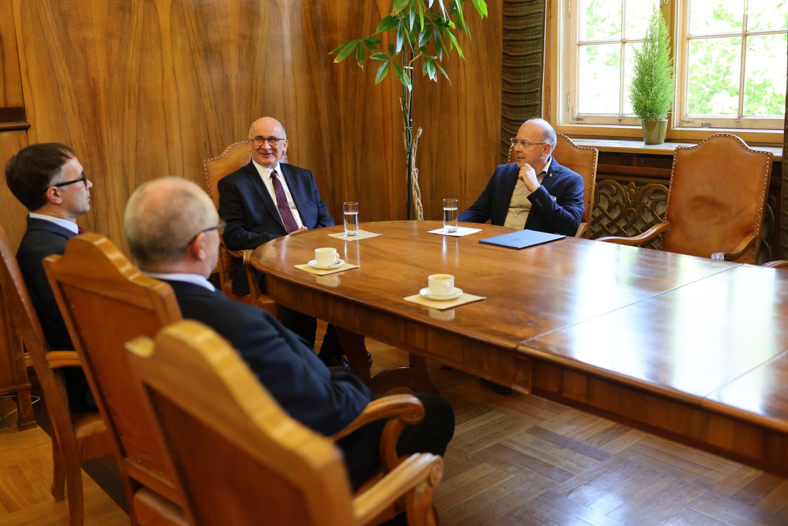 Grupa osób siedzi przy dużym drewnianym stole w eleganckiej sali i rozmawia podczas spotkania, na stole znajdują się filiżanki i szklanki z wodą.