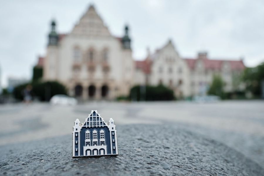 Zbliżenie na przypinkę w kształcie stylizowanego budynku Auli UAM ustawioną na kamiennej nawierzchni; w tle widoczny jest rozmyty gmach uczelni.