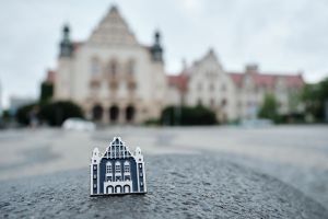Zbliżenie na przypinkę w kształcie stylizowanego budynku Auli UAM ustawioną na kamiennej nawierzchni; w tle widoczny jest rozmyty gmach uczelni.