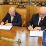 Dwie osoby siedzą przy drewnianym biurku i podpisują dokumenty podczas oficjalnego spotkania; na pierwszym planie widoczny jest stojak z oznaczeniem UAM.