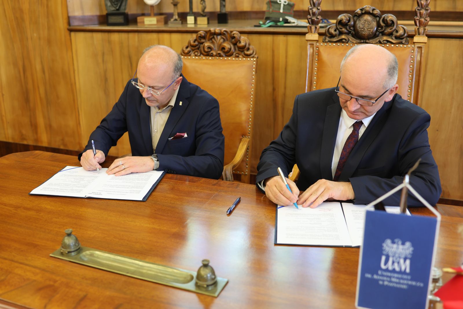 Dwie osoby siedzą przy drewnianym biurku i podpisują dokumenty podczas oficjalnego spotkania; na pierwszym planie widoczny jest stojak z oznaczeniem UAM.