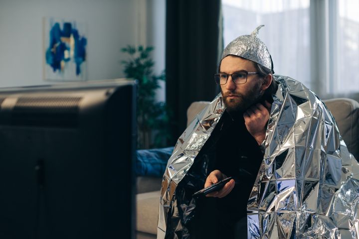Mężczyzna siedzi w salonie przed telewizorem, owinięty folią aluminiową i w czapeczce z folii, trzymając pilot w dłoni.
