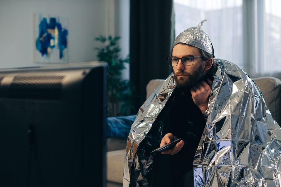 Mężczyzna siedzi w salonie przed telewizorem, owinięty folią aluminiową i w czapeczce z folii, trzymając pilot w dłoni.