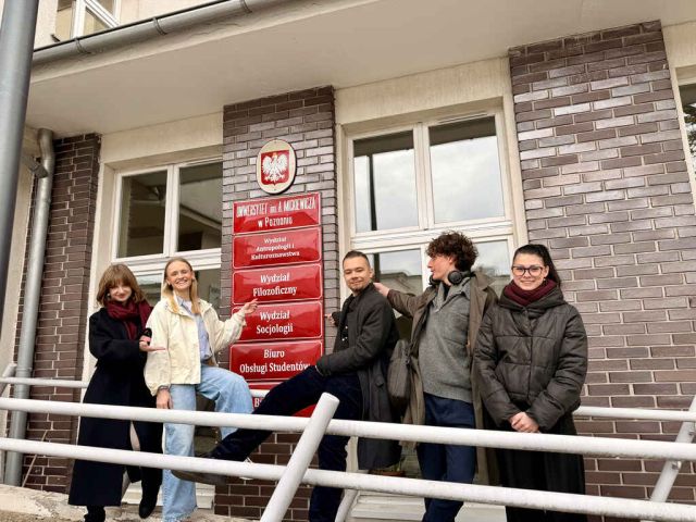 Pięcioro studentów przed budynkiem UAM w Poznaniu wskazuje tablicę informacyjną wydziałów