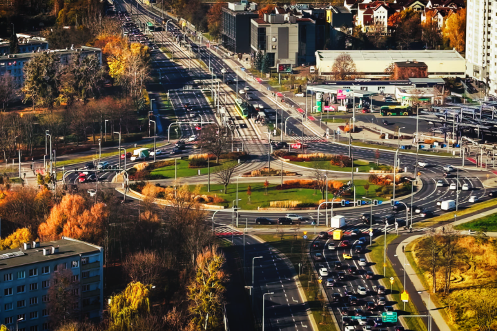 Widok z góry na duże rondo i wielopasmowe ulice z ruchem samochodowym w miejskiej zabudowie, otoczone jesiennymi drzewami.