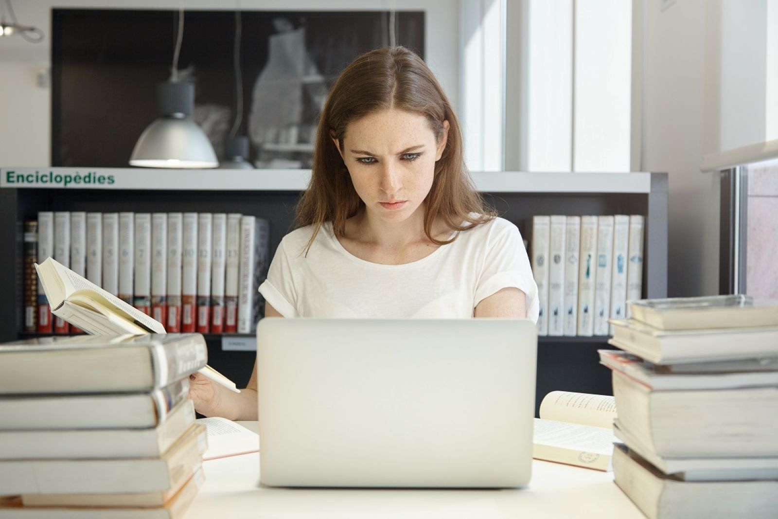 Kobieta w białej bluzce pracuje na laptopie w bibliotece, otoczona książkami. W tle regał z encyklopediami i napis „Enciclopèdies”.