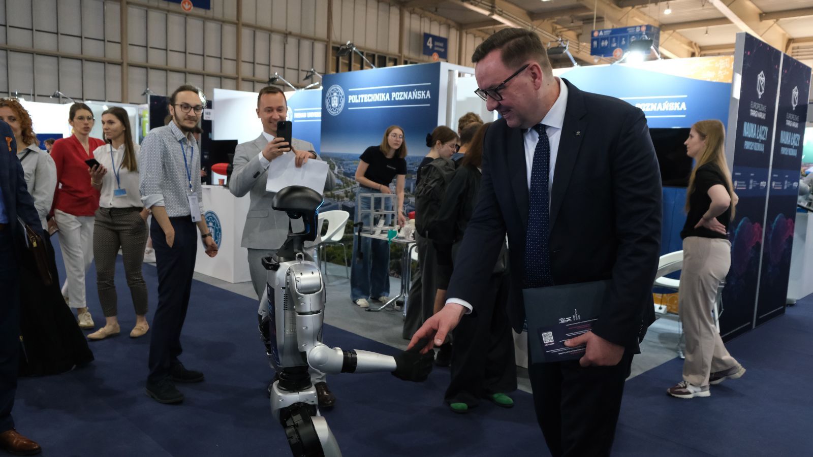 Osoba podaje rękę humanoidalnemu robotowi podczas targów nauki; wokół stoją uczestnicy wydarzenia, a w tle widoczne są stoiska wystawiennicze, w tym Politechniki Poznańskiej.