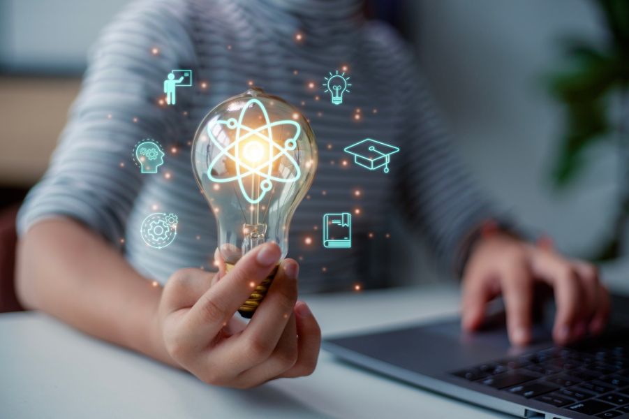 Osoba trzymająca żarówkę symbolizującą pomysł lub innowację, siedząca przy laptopie. Wokół żarówki unoszą się graficzne ikony związane z nauką i edukacją – atom, głowa z mózgiem, zębatka, książka, żarówka i czapka absolwenta.